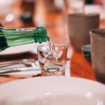 A hand pouring soju from a green bottle into a small glass at a Korean dinner table