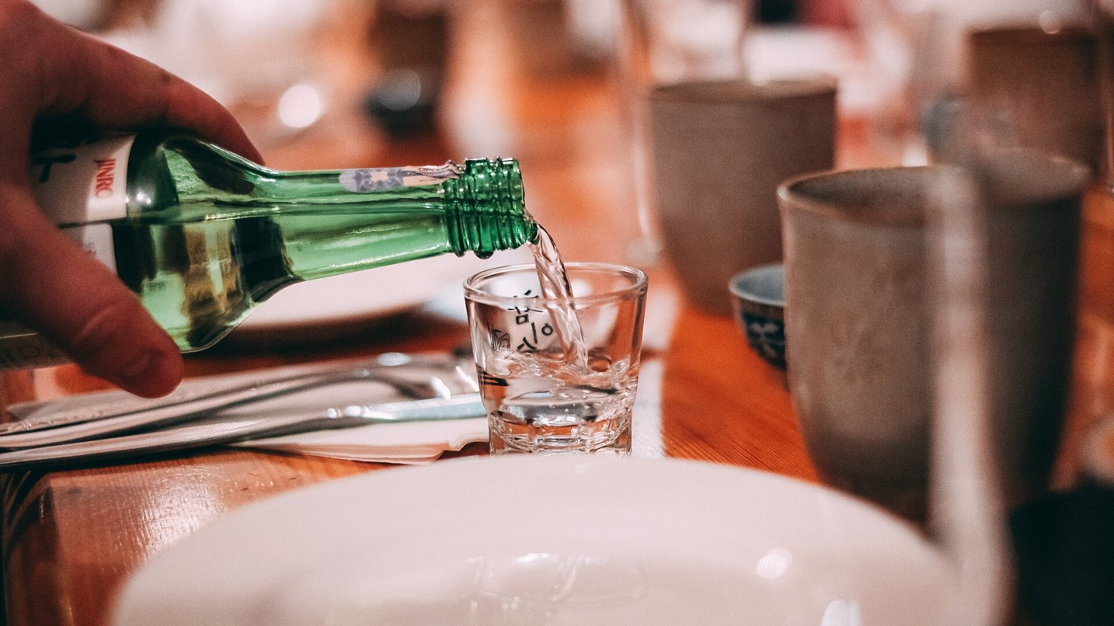 A hand pouring soju from a green bottle into a small glass at a Korean dinner table