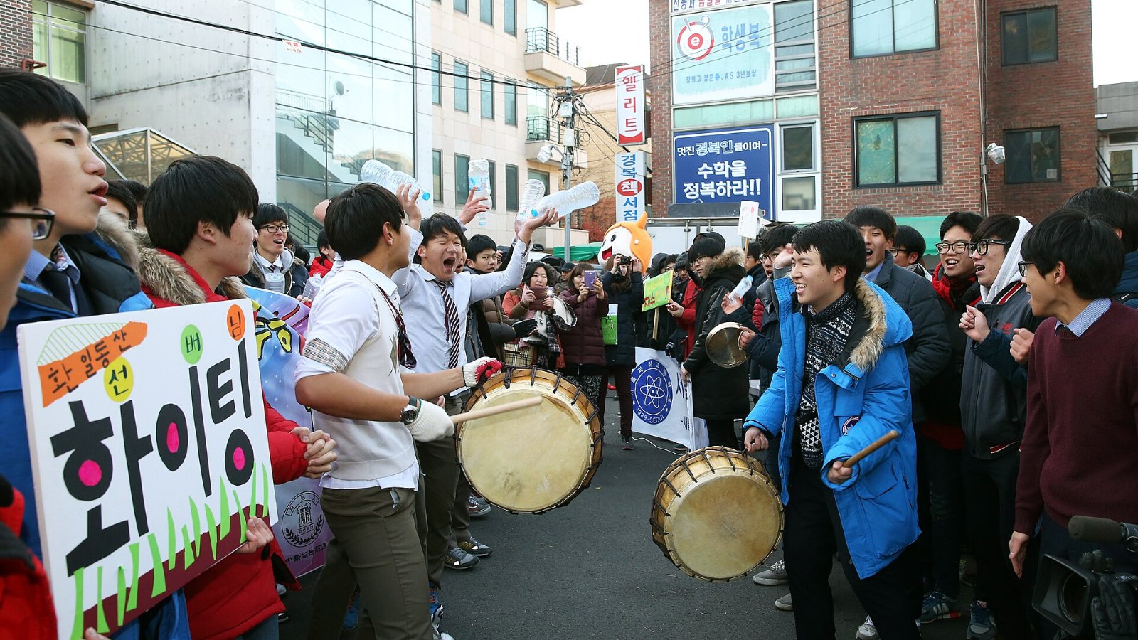 Students beating drums and holding a sign reading hwaiting outside a Suneung test site